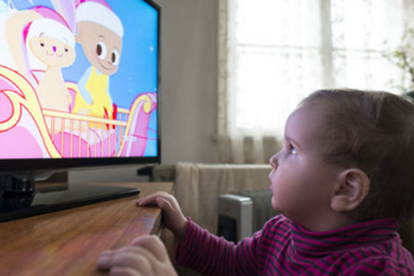 Маленькая девочка стоит возле экрана и смотрит мультик